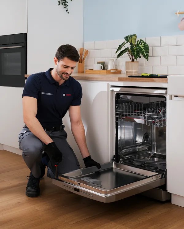 Técnico joven sonriente instalando o reparando lavavajillas integrado en cocina blanca moderna