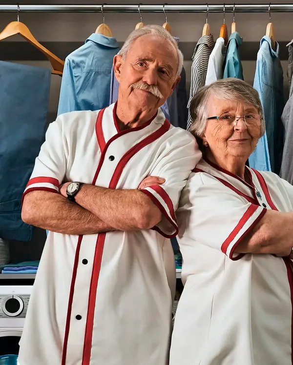 Pareja mayor en uniforme blanco con rayas rojas posando frente a perchero con ropa y electrodomésticos